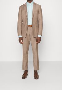 Costume marrone chiaro con una vestibilità su misura, abbinato a una camicia azzurra. Una cintura e scarpe in pelle marrone completano l'outfit.