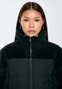 Černá zipová bunda s fleecovou vrchní částí a kontrastní hladkou látkou dole. Vysoký límec a boční kapsy dotvářejí design.