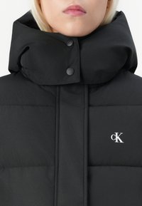 Sort puffervest med høy krage, med trykknapper og en hvit logo på brystet. Glatt, quiltet tekstur med horisontale segmenter.