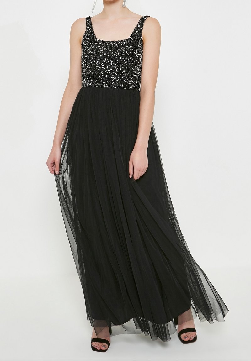 Femme portant une robe de soirée noire sans manches avec un corsage à sequins et une jupe fluide en tulle, associée à des talons noirs ouverts.