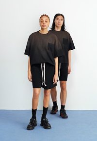 Zwarte oversized t-shirts met borstzakken, gecombineerd met zwarte shorts met witte koordjes. Modellen dragen dikke zwarte schoenen en zwarte sokken.