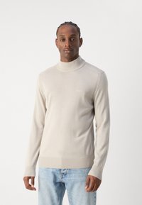 Suéter de cuello alto de color beige claro, hecho de una tela suave con puños y dobladillo acanalados, que presenta un sutil detalle del logo en el pecho.