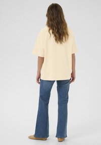 T-shirt à manches courtes, de couleur jaune pâle, avec une coupe ample, associé à un jean évasé bleu et des chaussures légères de couleur marron. Le tissu semble doux.