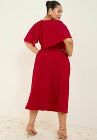 Robe midi rouge avec des manches flottantes, fermeture à nouer dans le dos, tissu lisse et talons avec lanières à la cheville sur un sol texturé.