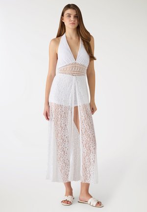 Kobieta w białej koronkowej sukience na szyję bez rękawów z przezroczystymi panelami i przednim rozcięciem, w połączeniu z białymi płaskimi sandałami.