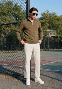 Homme portant des lunettes de soleil, un pull vert olive, un pantalon blanc et des baskets blanches, debout près d'une clôture près d'un terrain de basket en plein air par une journée ensoleillée.