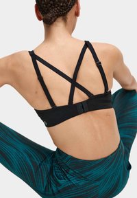 Zwarte sportbeha met kruislings straps en verstelbare clips. Het model draagt een patroon van teal leggings met zwarte accenten.