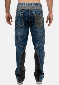Jeans en denim bleus avec des sections à délavage clair et foncé, dotés d'une poche arrière en camouflage et d'une couture contrastante. Design à jambe fuselée.