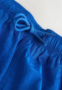 Gros plan sur un short de bain bleu à motif géométrique avec un cordon tressé noué en boucle à la ceinture élastique.