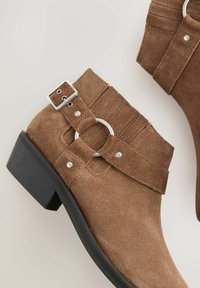 Botines de ante marrón con una hebilla decorativa y un anillo de metal, con un tacón bajo negro y detalles de costura en la parte superior.