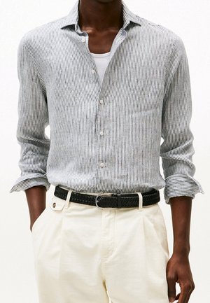 Homme portant une chemise boutonnée à rayures verticales gris clair sur un sous-vêtement blanc, associée à un pantalon crème et une ceinture noire tissée.
