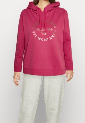 Vrouw die een roze Tommy Hilfiger hoodie draagt met gedrukte tekst en lichtgrijze joggingbroek, staand tegen een effen achtergrond.