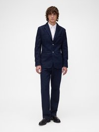 Σκούρο τζιν κοστούμι με σακάκι με πέταλο γιακά και παντελόνια ίσιας γραμμής. Λευκό πουκάμισο με κουμπιά από κάτω. Γυαλιστερά μαύρα παπούτσια.