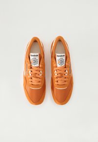 Reebok oransje joggesko med en glatt tekstiloverdel, hvite detaljer og en flat gummisåle. Har rund tå og snøring.