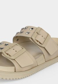 Μπεζ σανδάλι με δύο ρυθμιζόμενους ιμάντες με αγκράφες, κάθε ένας διακοσμημένος με ασημένια καρφιά σε σχήμα πυραμίδας και υφασμένο πέλμα για καλύτερη πρόσφυση.