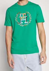 Camiseta verde de algodón con un logo bordado multicolor prominente en el centro, rodeado por un diseño de rama de olivo. Mangas cortas.
