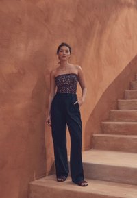 Stropfri mønstret top i mørke nuancer, kombineret med sorte bukser med høj talje. Modellen står på rustikke trin mod en okkerfarvet væg.