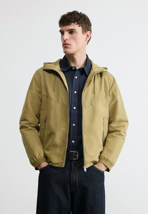 Bărbat tânăr purtând o jachetă cu glugă de culoare cămilă peste o cămașă din denim închis cu nasturi și blugi închiși la culoare, cu mâinile în buzunarele jachetei, uitându-se spre stânga.