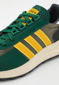 Grön och gul sneaker med strukturerade material, tre gula ränder och en tjock vit mellansula med ett mönstrat ytskikt.