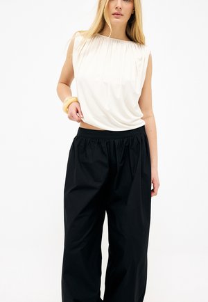 Femme blonde portant un haut blanc sans manches et un pantalon noir ample, posant avec une main touchant son haut et portant un bracelet jaune.