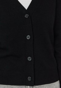 Cardigan en maille noire avec encolure en V et quatre boutons foncés à l'avant, porté sur un haut blanc.