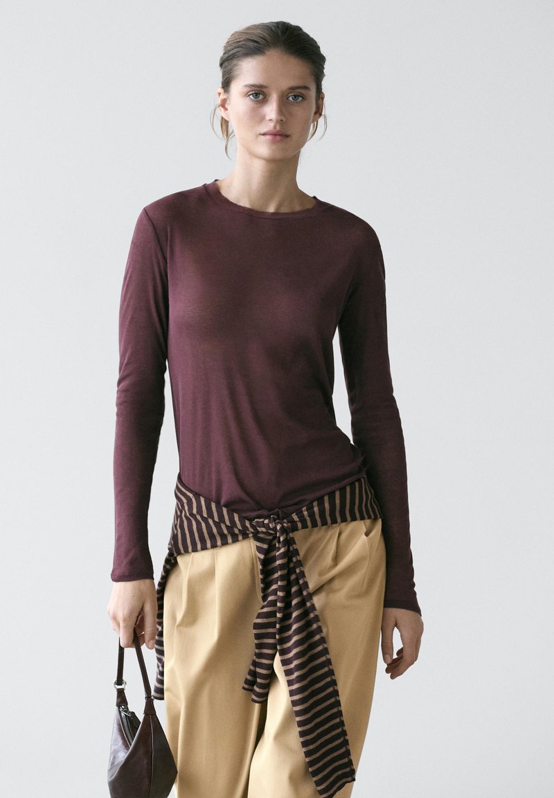 Jeune femme portant une chemise à manches longues bordeaux, un pantalon beige, avec un pull rayé attaché autour de la taille, tenant un sac à main foncé.