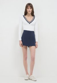 Frau in einem weißen Langarm-Oberteil mit V-Ausschnitt und Marineblau gestreift, Marineblau hochgeschnittenen Shorts und weißen Sneakers, stehend vor einem schlichten weißen Hintergrund.