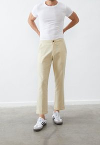 Pantalon beige clair, coupe droite, avec fermeture à bouton sur le devant, poches latérales et texture subtile. Porté avec un t-shirt blanc et des baskets.