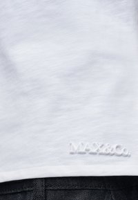 Tissu blanc avec un petit logo en relief « MAX&Co. » près de l'ourlet au-dessus d'un jean en denim foncé.