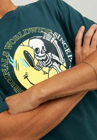 Camiseta de algodón en color verde azulado con un gráfico de un esqueleto y una cara sonriente sobre un fondo amarillo. Manga corta y corte relajado.