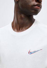 Κοντινό πλάνο στον λαιμό και το κάτω μέρος του προσώπου ενός ατόμου που φοράει λευκό πουκάμισο με ένα μικρό ιριδίζον λογότυπο Nike Swoosh στο στήθος.