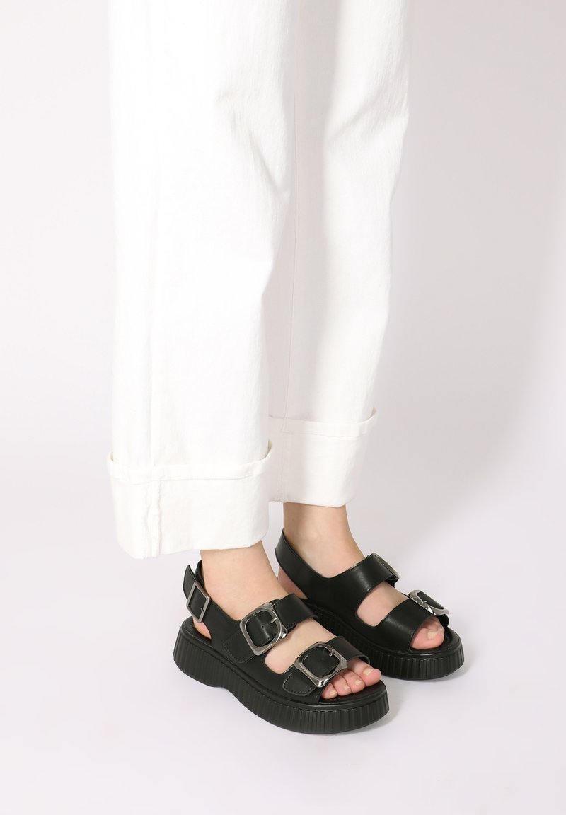 Zwarte sandalen met twee verstelbare gespen, stevige zool, open teenontwerp en soepel leren materiaal, gecombineerd met witte cropped broeken.