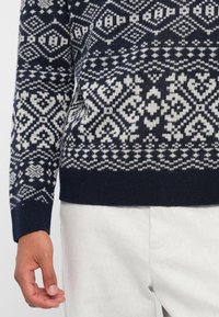 Granatowy sweter dzianinowy z białym geometrycznym wzorem. Ribbowane mankiety i dół, teksturowana tkanina, dopasowany krój. Połączony z jasnymi spodniami.