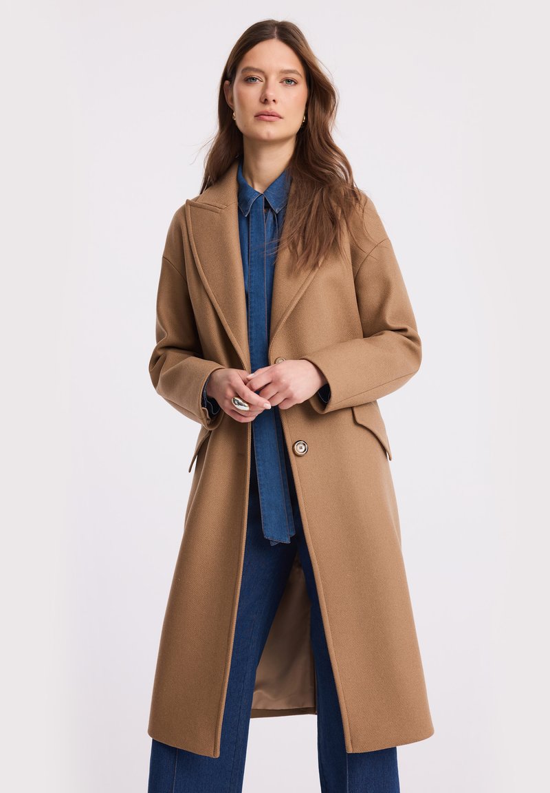 Młoda kobieta z długimi brązowymi włosami, ubrana w długi płaszcz w kolorze karmelowym, na niebieskiej koszuli dżinsowej i dżinsach, stoi z złożonymi dłońmi.