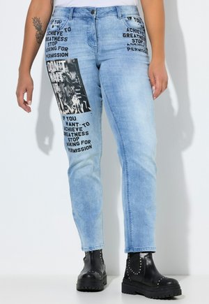 Lyseblå jeans med sort bloktekst og grafiske patches, båret med sorte ankelstøvler med nitter, tatovering synlig på venstre underarm.