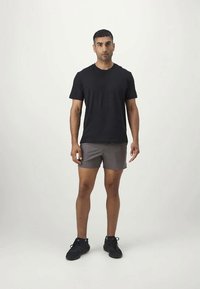 Crna pamučna majica, sivi šortsevi i crne atletske tene. Model stoji naslonjen na jednostavnu bijelu pozadinu, ruke opuštene uz tijelo.