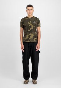 Roheline kamuflaažiga lühikeste varrukatega t-särk, mustad cargo püksid ja pruunid mustrilised tossud. Mudel seisab ühevärvilise valge tausta ees.