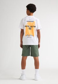 Witte katoenen T-shirt met oranje en zwarte grafische tekst op de achterkant, gecombineerd met groene shorts. Heeft een ronde hals en een relaxte pasvorm.