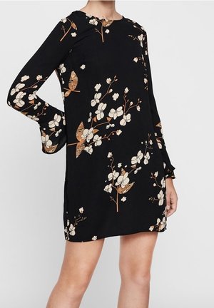 Czarna sukienka z długim rękawem, z kremowo-brązowymi kwiatowymi wzorami, z lekko rozszerzanymi mankietami, noszona przez osobę, która jedną ręką opiera się na biodrze.