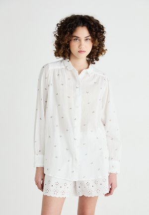 Weißes Hemd mit knöpfbarem Kragen, mit einem strukturierten, gestreiften Muster und kleiner floralem Stickerei. Kombiniert mit weißen Shorts mit gewelltem Saum.