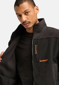 Giacca in pile nera con colletto alto, dettagli arancioni della zip e un logo. Presenta un mix di tessuto testurizzato e pannelli lisci.