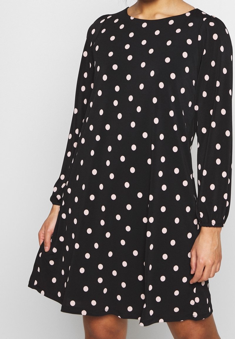 Femme portant une robe noire à manches longues avec des pois rose clair, debout les mains détendues le long du corps.