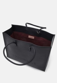 Sac fourre-tout en cuir noir avec une texture granuleuse, ouverture sur le dessus, deux poignées, et un intérieur bordeaux avec une petite poche latérale.