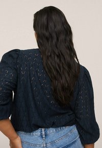 Femme aux longs cheveux ondulés et foncés portant un haut bleu marine à œillets et un jean taille haute bleu clair, vue de dos.