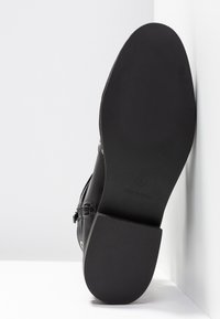 Semelle de botte noire avec talon texturé et avant lisse, debout verticalement contre un fond blanc en angle, taille 4, fabriquée en Chine.