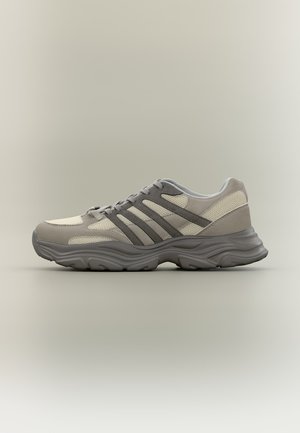 Zapatilla deportiva gris y beige con paneles de malla, superposiciones de ante y suela gruesa con dibujo, mostrada de perfil sobre un fondo neutro.