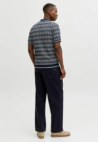 Man standing facing sideways wearing a patterned short-sleeve shirt, dark wide-leg pants, and light brown shoes on a plain background.