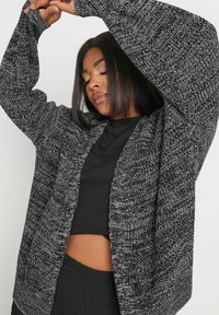 Stickad kofta i svart och vit melerad mönster, med breda ärmar och öppen front. Mjuk textur och avslappnad passform.