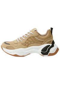 Chaussure de sport beige avec une tige texturée, des lacets blancs et une semelle épaisse blanche et gomme. Présente des accents noirs sur le talon et le côté.