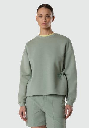 Femme portant un haut à manches longues vert sauge avec des liens latéraux ajustables et un short vert assorti, debout devant un fond clair.